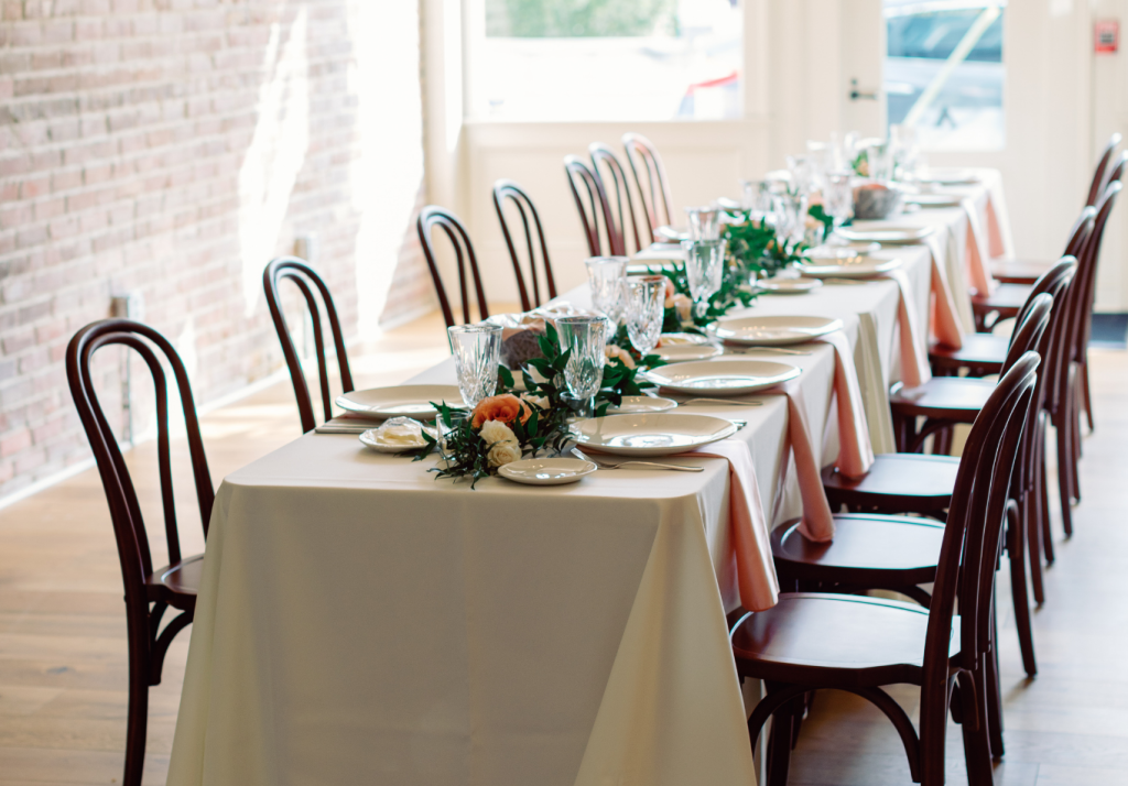A long dining table set for a meal with white tablecloths, glassware, plates, and floral centerpieces, surrounded by wooden chairs in a sunlit room with brick and white walls at The Rutledge Room in Charleston.