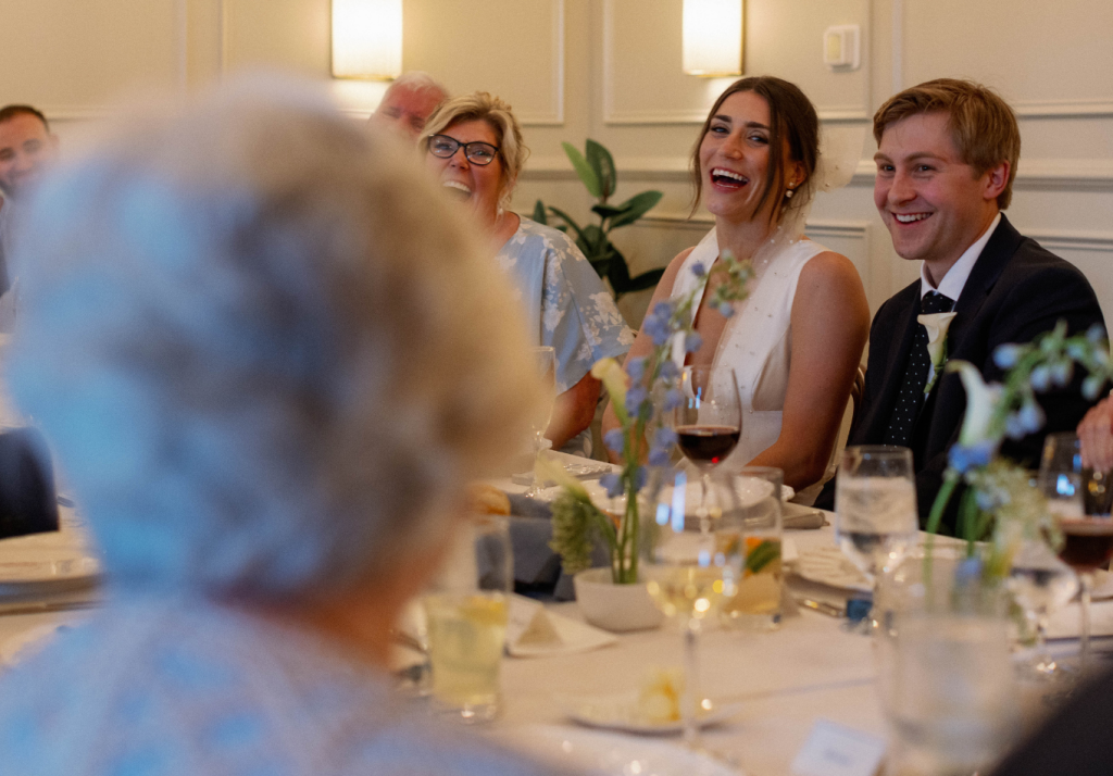 A bride and groom sit at a decorated table, smiling and laughing with guests during a wedding reception. Drinks and flowers are on the table, and the atmosphere is warm and joyful.