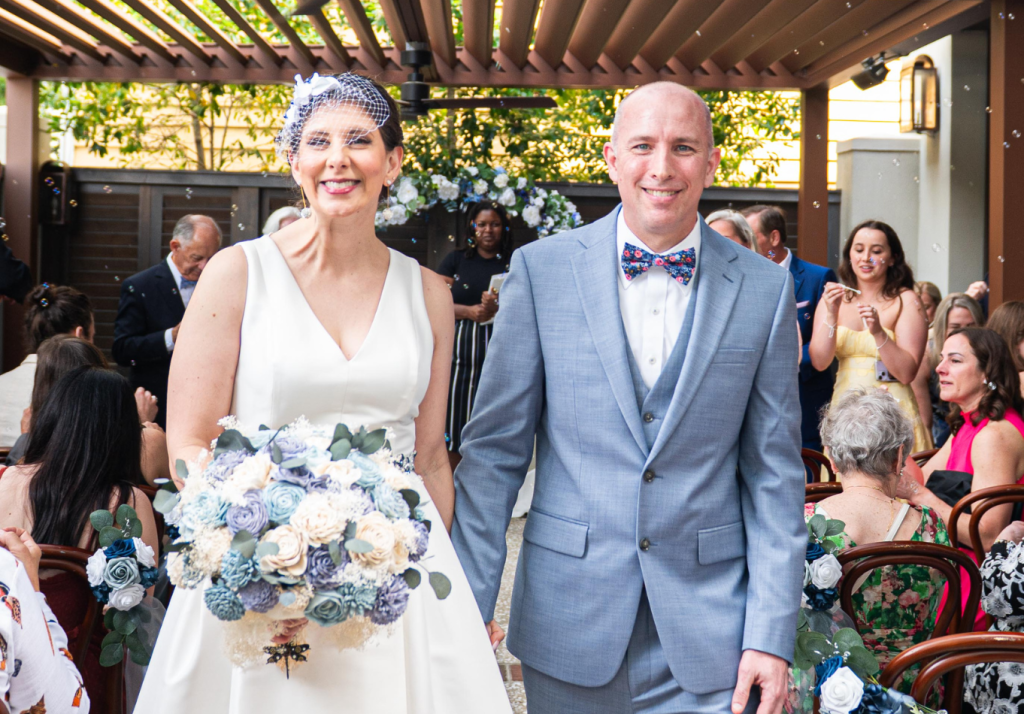 A smiling bride in a white dress and a groom in a light blue suit with a bow tie walk down the aisle outdoors, surrounded by seated guests and floral decorations. The bride holds a large bouquet.