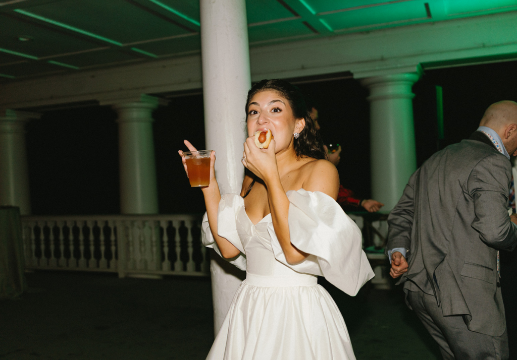 A woman in a white off-shoulder wedding dress eats a hot dog while holding a drink, standing next to a white column at a wedding reception. Other guests in formal wear are in the background.