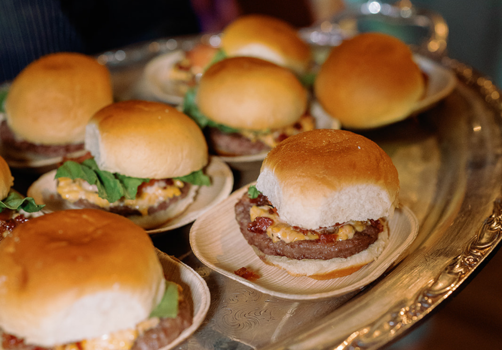 A close-up of several small beef sliders with lettuce and cheese on round buns, arranged on a silver tray.