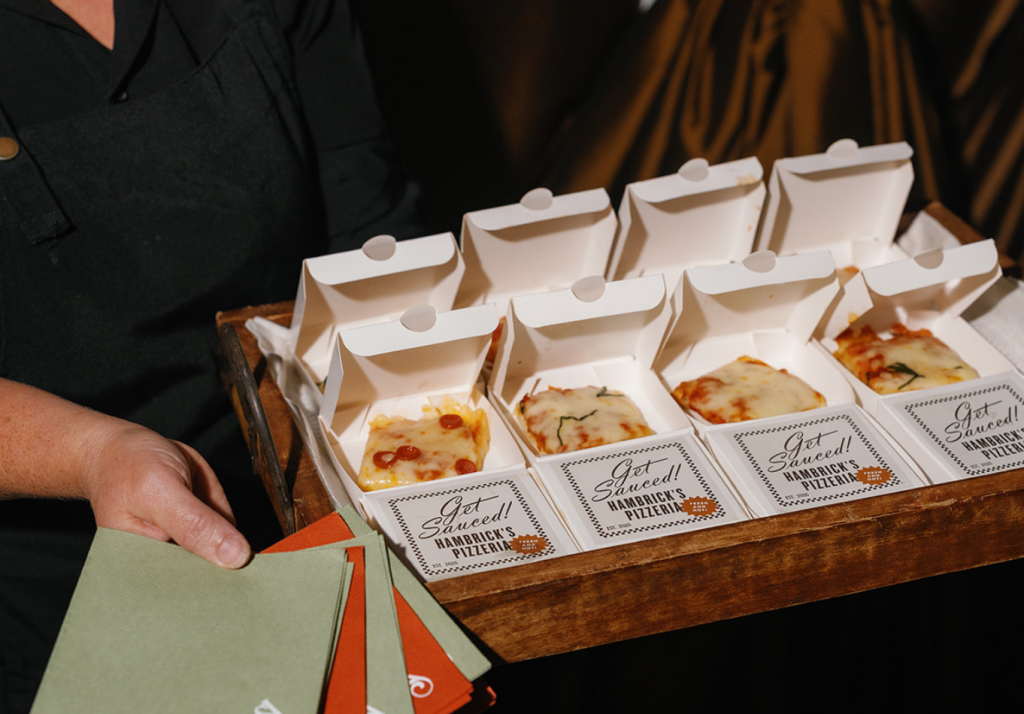 A server holds a wooden tray with several small pizzas in individual white boxes, labeled Get Sauced! Green and red napkins are visible in their other hand.