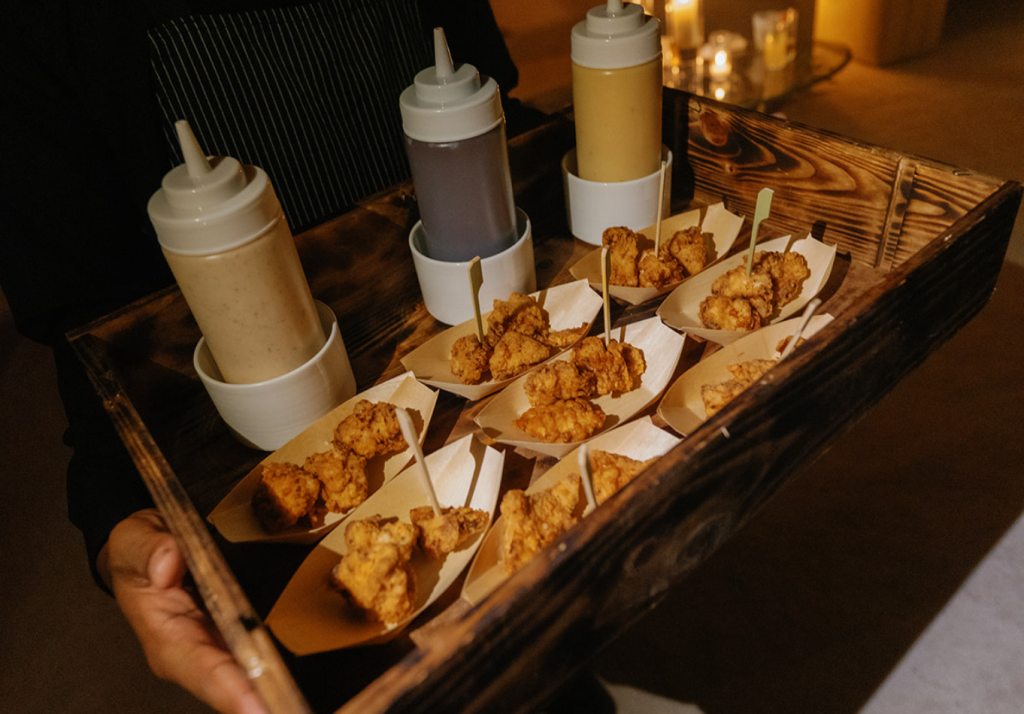 A wooden tray holds several small paper trays filled with fried chicken nuggets and wooden picks, along with three squeeze bottles of different sauces. The background is softly lit with candles.
