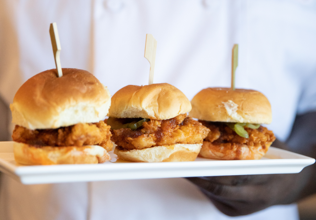 Three fried chicken sliders with buns, each held together with a wooden pick, are arranged on a white rectangular plate held by a chef in a white shirt.
