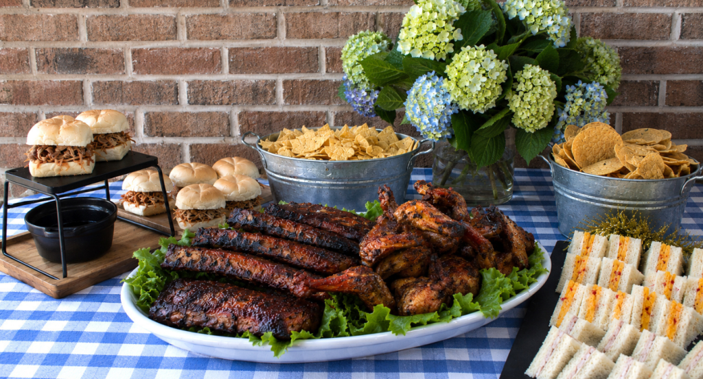 Hamby Catering tailgate spread with barbecue ribs, chicken, sliders, tea sandwiches and chips on a blue checkered table in Charleston, SC.