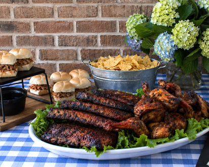 Hamby Catering tailgate spread with barbecue ribs, chicken, sliders, tea sandwiches and chips on a blue checkered table in Charleston, SC.
